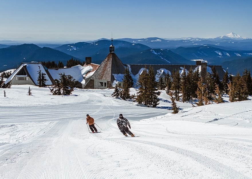 TWO SKIERS HEADING TOWARDS THE BACK OF TIMBERLINE LODGE ON MT. HOOD WITH THE CASCADE RANGE AND MT JEFFERSON IN THE BACKGROUND