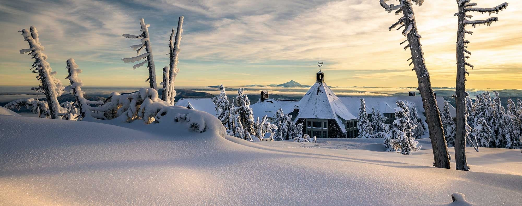 Timberline Lodge pictured from a snowy bank at sunset with Mt. Jefferson in the background