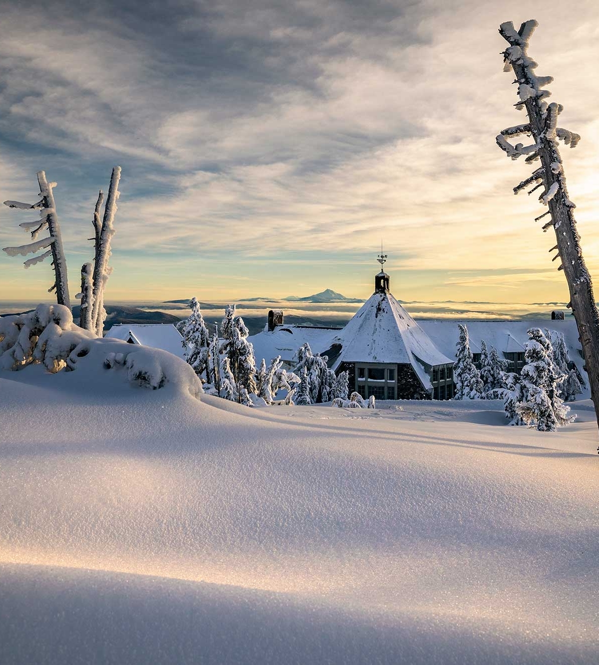 Timberline Lodge pictured from a snowy bank at sunset with Mt. Jefferson in the background