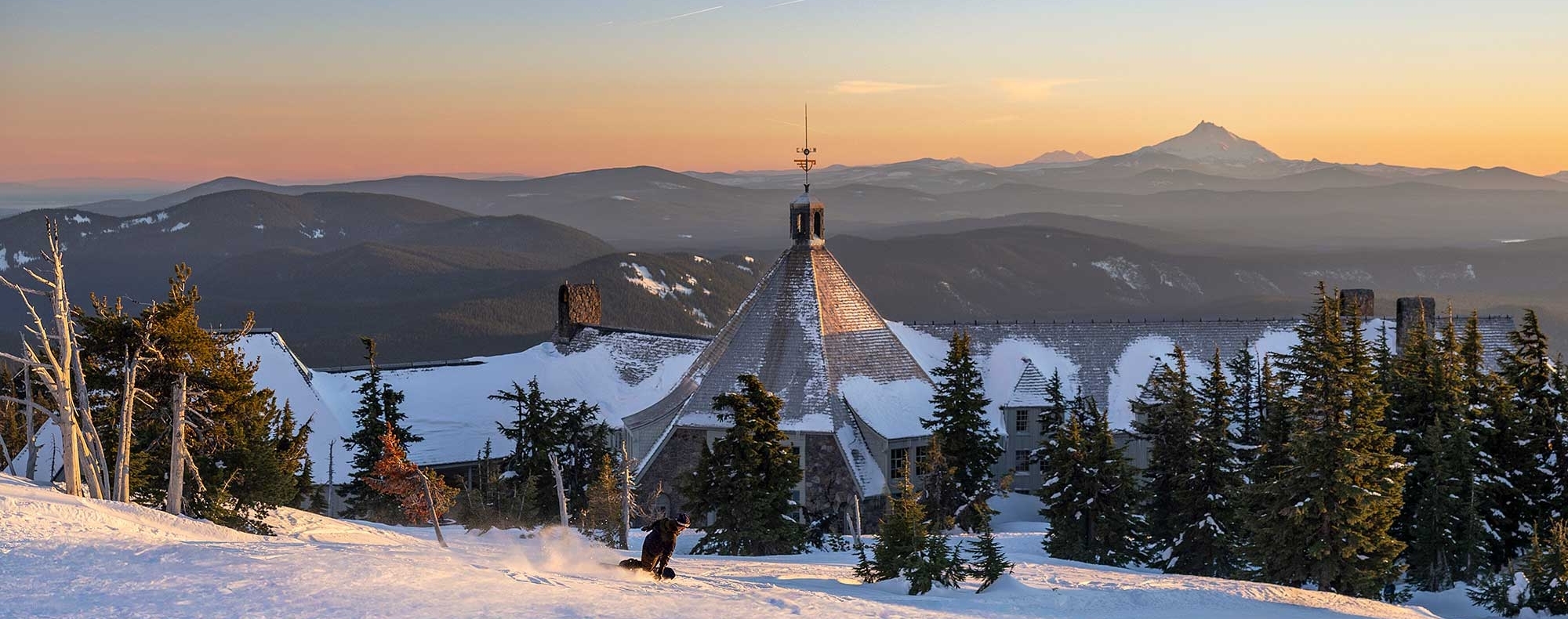 Snowboarder cruising down the slopes into the back of Timberline Lodge at sunset