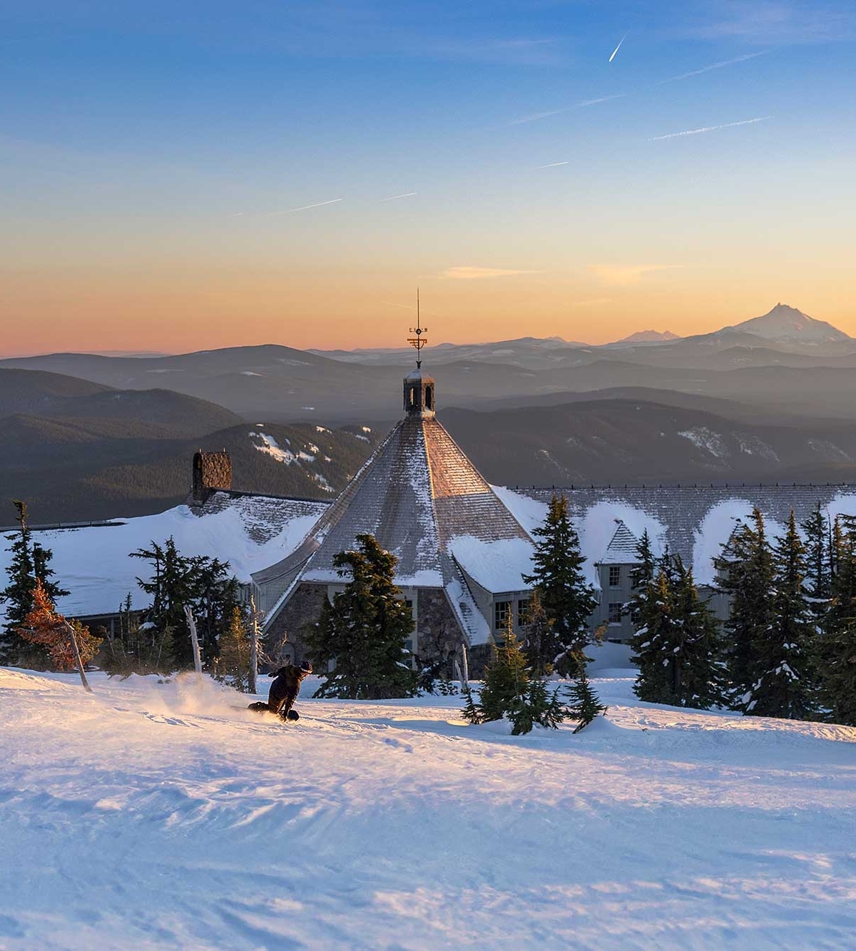 Snowboarder cruising down the slopes into the back of Timberline Lodge at sunset