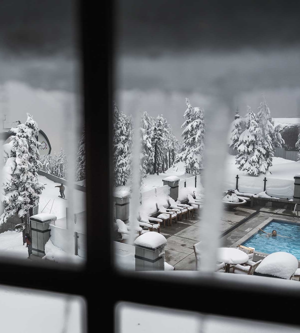 View through a frosted window of the Timberline Lodge pool area, surrounded by snow-covered trees and furniture, with a person swimming in the heated outdoor pool amidst a winter landscape.