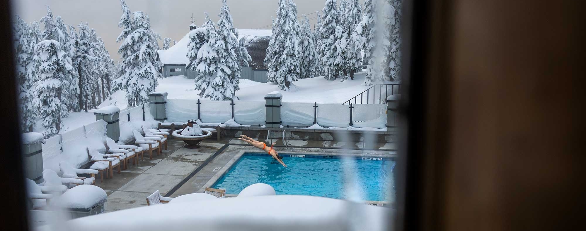 View through a frosted window of the Timberline Lodge pool area, surrounded by snow-covered trees and furniture, with a person swimming in the heated outdoor pool amidst a winter landscape.