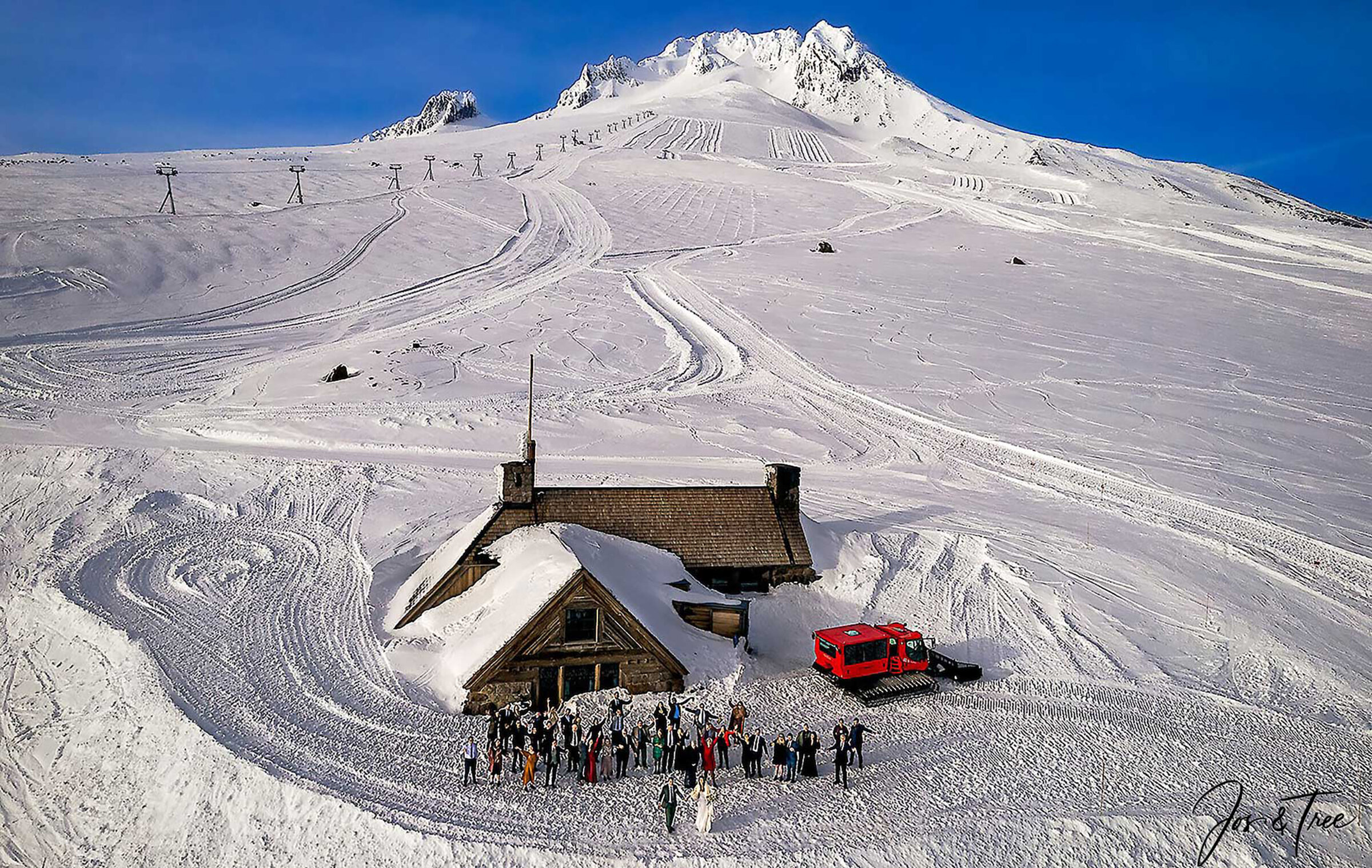 An aerial view of Silcox Hut on Mt. Hood, surrounded by deep snow, with a group of people gathered outside and a red snowcat parked nearby. Freshly groomed ski tracks cover the mountain slopes under a clear blue sky.