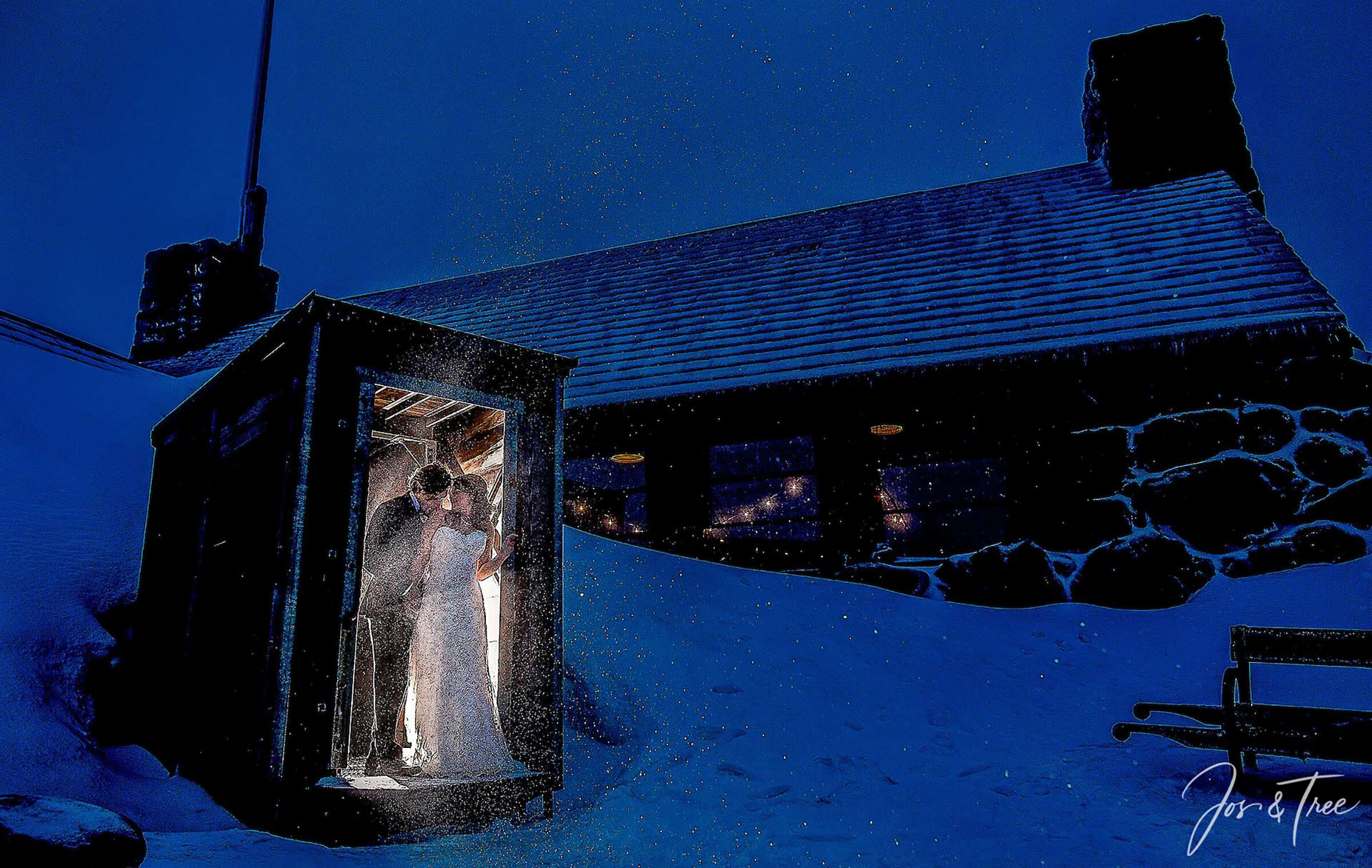 A bride and groom share a romantic kiss in the softly lit entryway of Silcox Hut at night, surrounded by falling snow and the dark, snow-covered stone exterior of the historic mountain lodge.