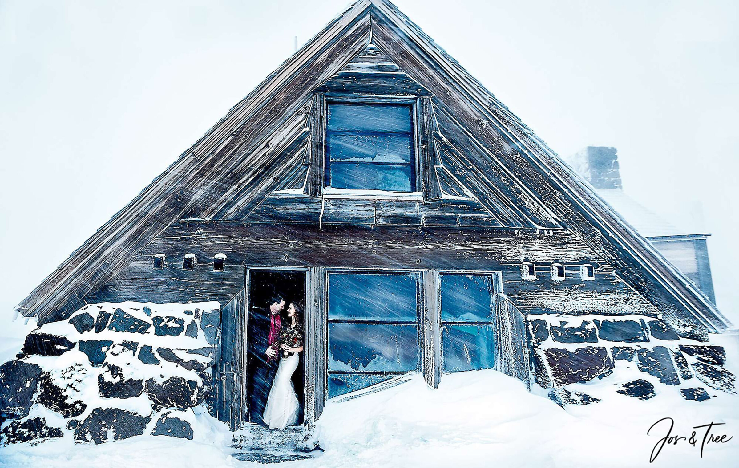 A bride and groom stand in the doorway of Silcox Hut during a snowy winter storm, framed by the rustic wooden exterior and stone foundation of the historic mountain lodge as wind sweeps snow across the scene.