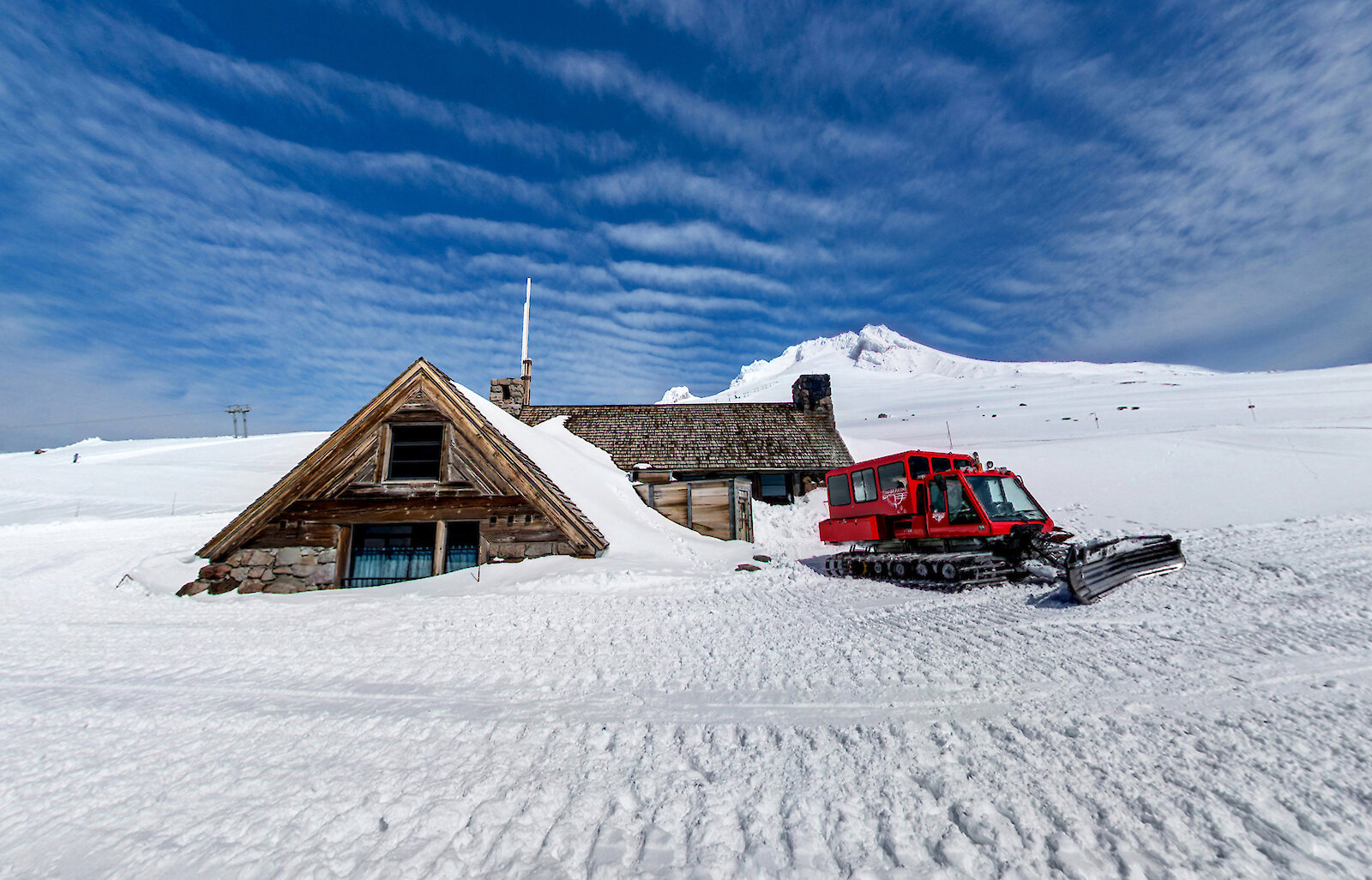 Snow-covered Silcox Hut above Timberline Lodge with a bright red snowcat parked beside it under a blue sky.