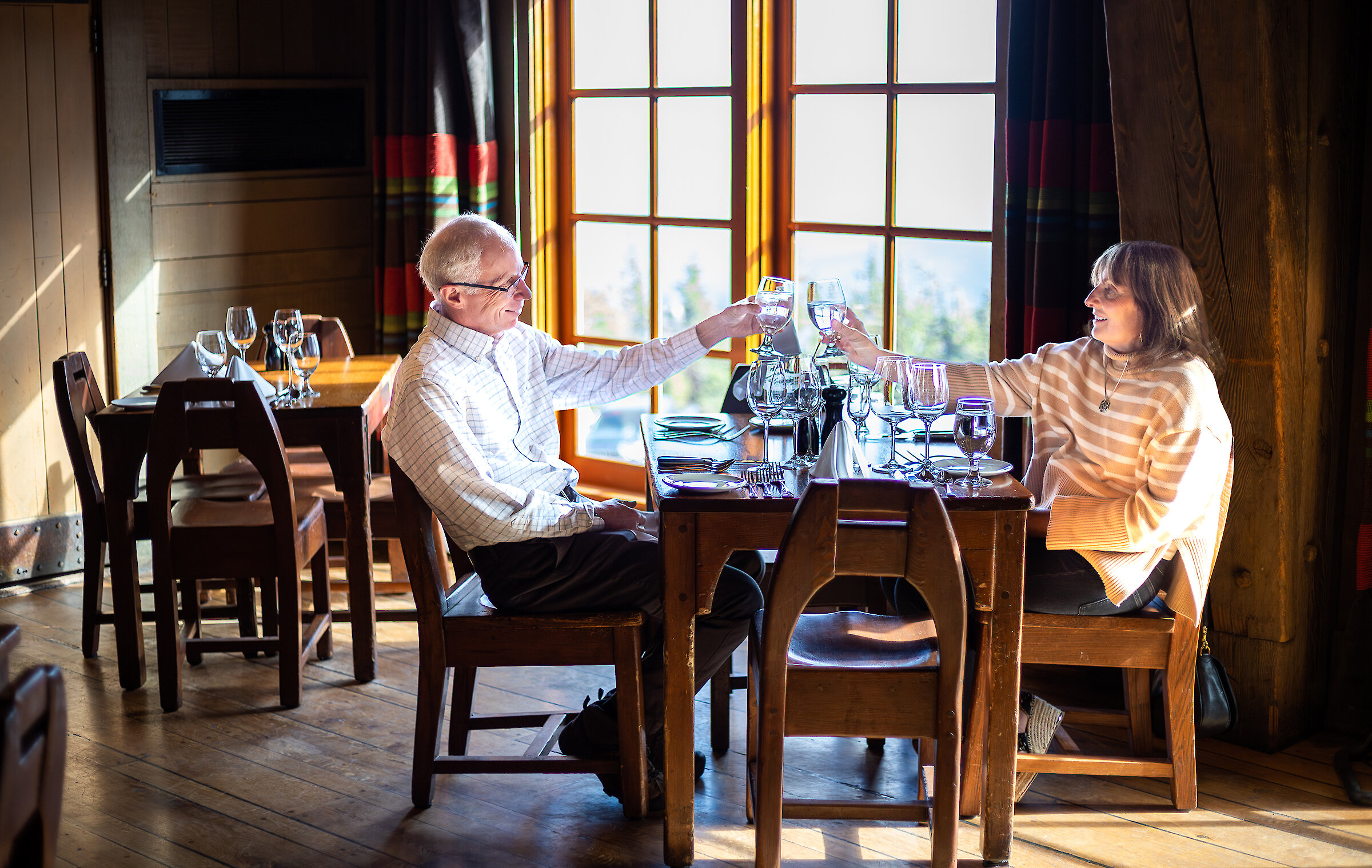 Couple toasting with wine at a sunlit dining table inside Timberline Lodge.