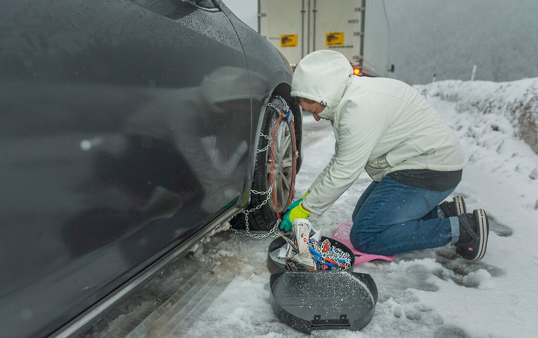 A person kneels on a snowy roadside, fastening tire chains onto a car’s wheel while snow and slush cover the ground. A chain case sits open nearby, and a large truck is parked ahead in wintry, low-visibility conditions.