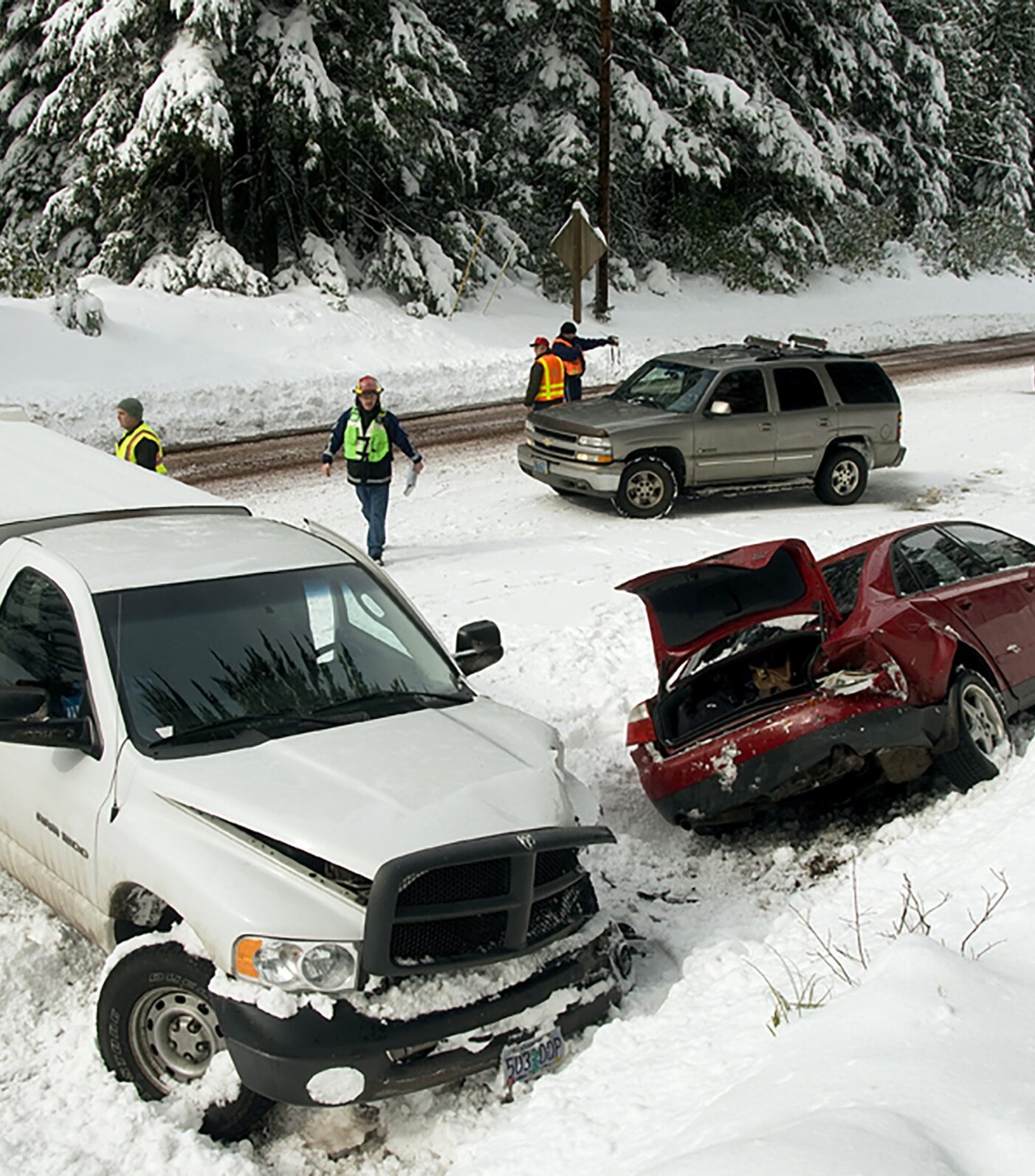 Two damaged vehicles sit in a snowy roadside ditch after a winter crash, with emergency responders and law enforcement officers on scene. Snow-covered trees and passing traffic line the highway as crews work in hazardous conditions.