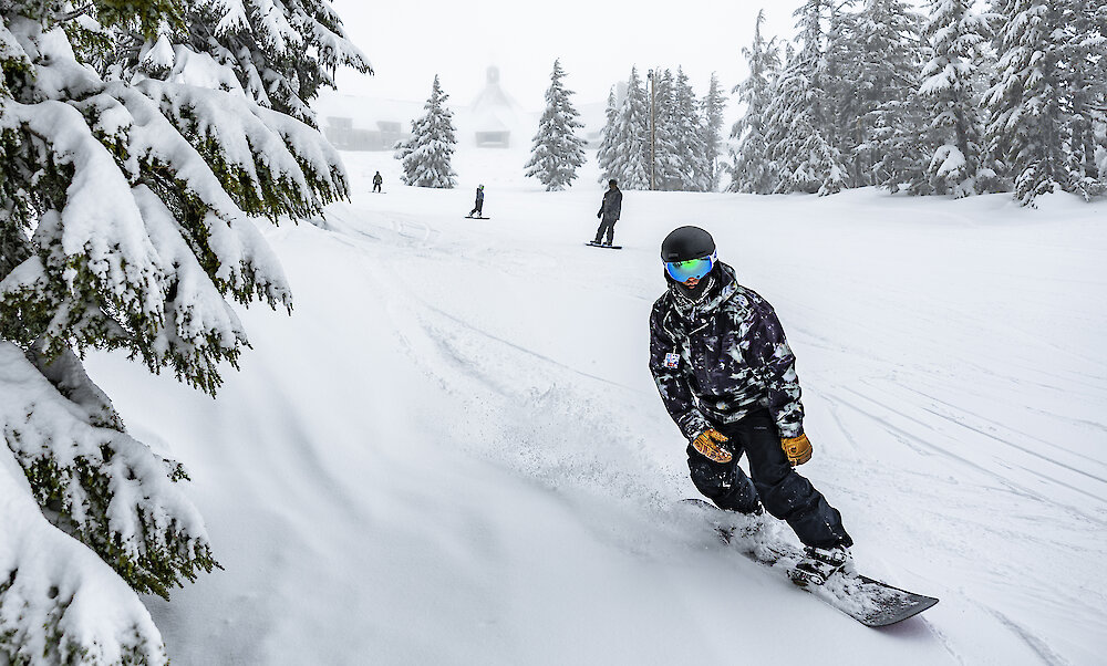 A snowboarder in a helmet and mirrored goggles carves through fresh powder on a wide, snowy run, with snow-laden evergreens and a few riders fading into the foggy background.