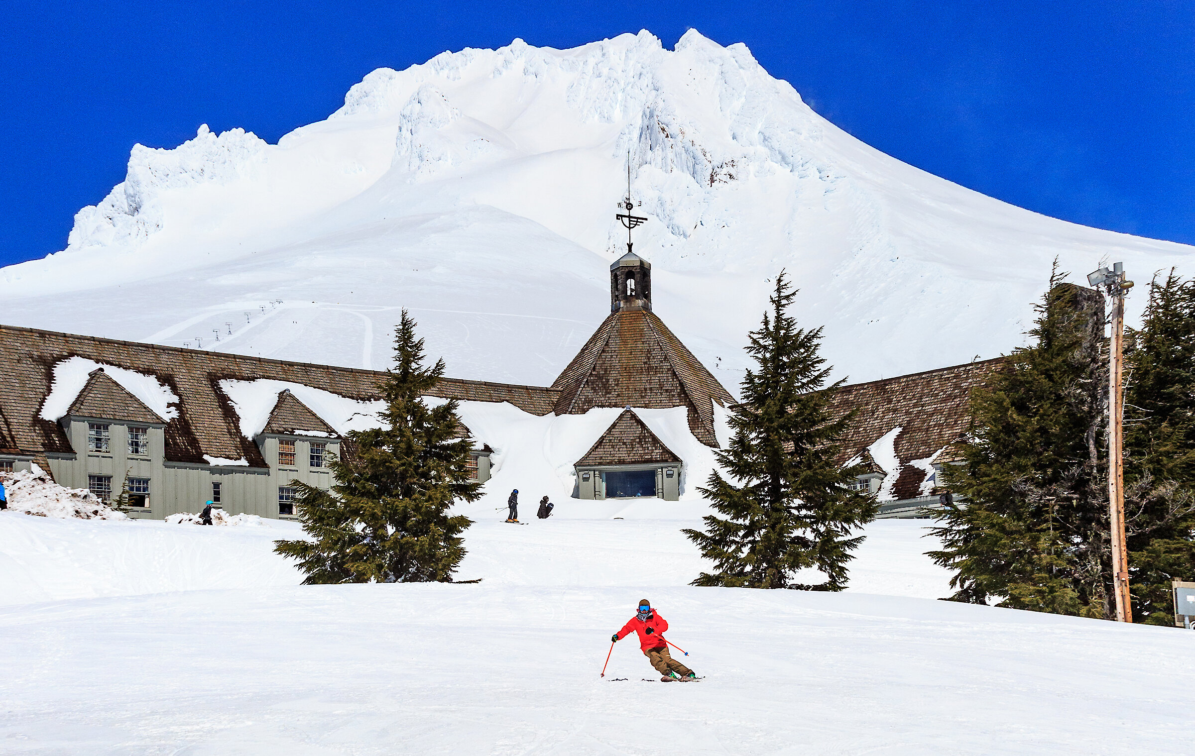 Skier in a red jacket carves across a snowy slope in front of Timberline Lodge, with evergreen trees and the sunlit, glacier-covered Mt. Hood rising under a deep blue sky.