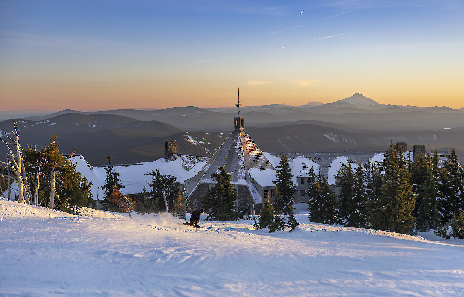 Sunset view over a snowy ski slope at Timberline Lodge. The lodge’s steep, pyramid-shaped roof rises above evergreen trees while a lone skier carves down the foreground. Layers of dark mountain ridges stretch into the distance under a pastel orange-to-blue sky.