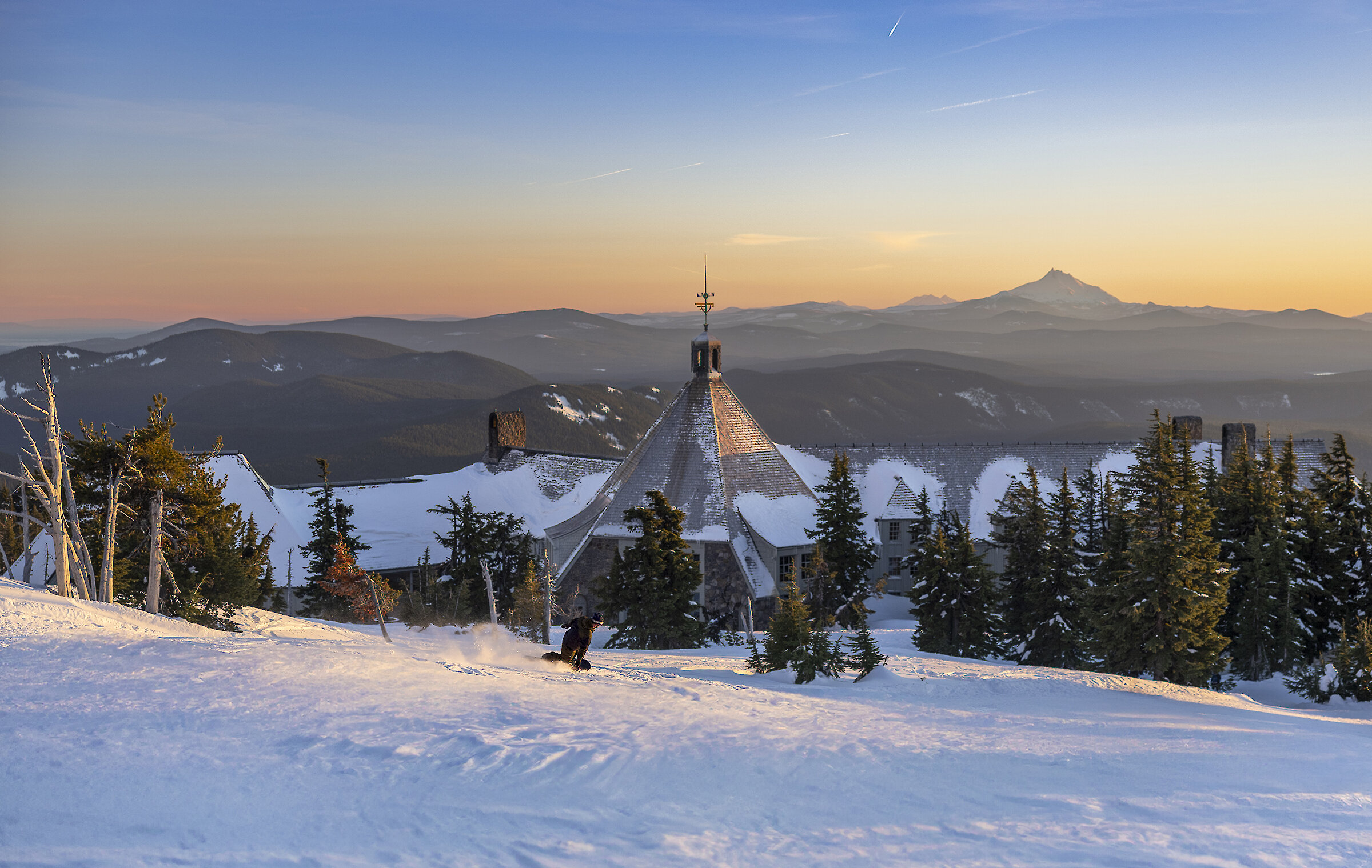 Sunset view over a snowy ski slope at Timberline Lodge. The lodge’s steep, pyramid-shaped roof rises above evergreen trees while a lone skier carves down the foreground. Layers of dark mountain ridges stretch into the distance under a pastel orange-to-blue sky.