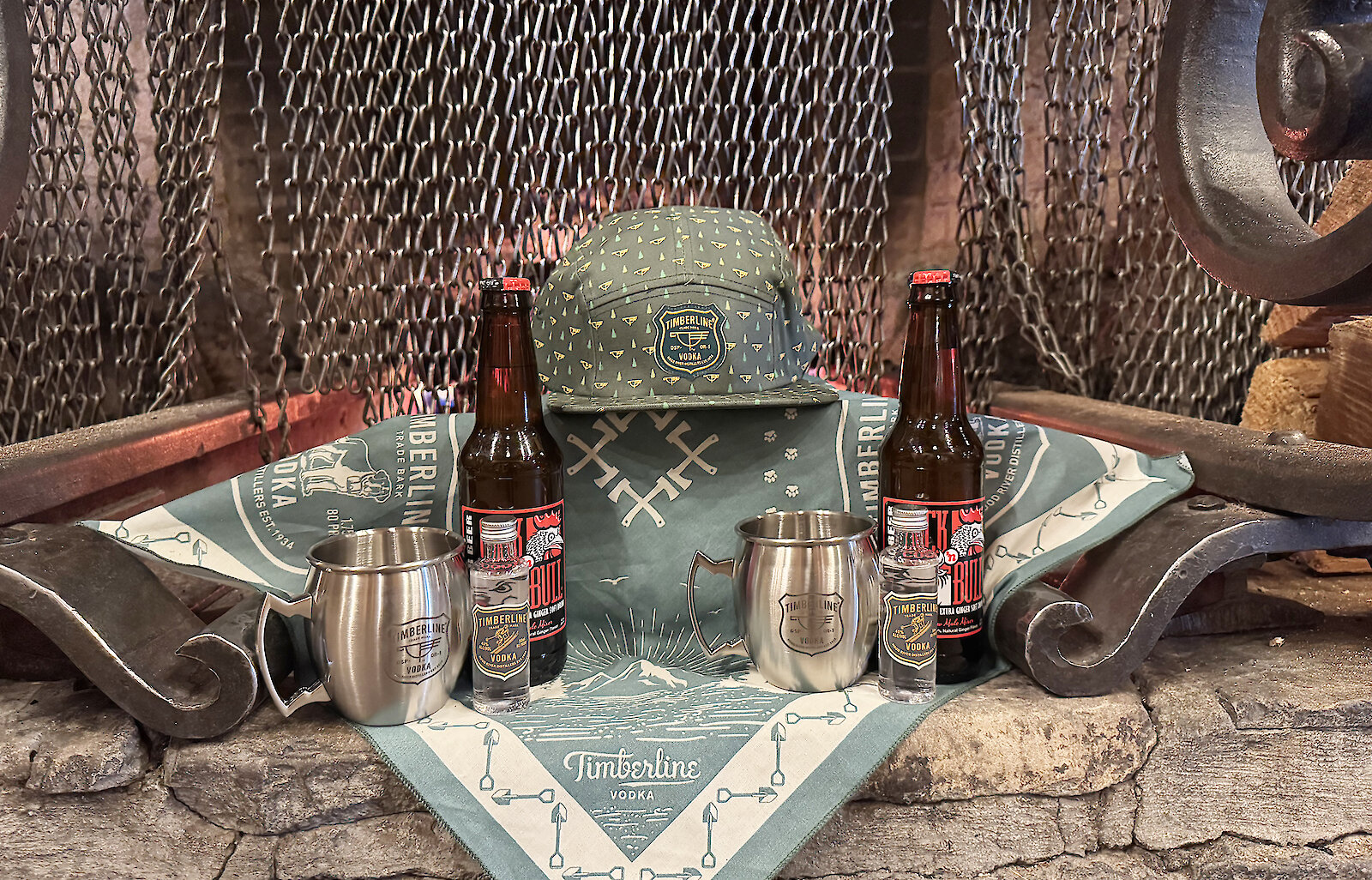 Timberline Vodka branded merchandise displayed by a fireplace, including stainless steel mule mugs, bottled vodka, craft beer, a green Timberline hat, and a branded bandana arranged on a stone hearth.