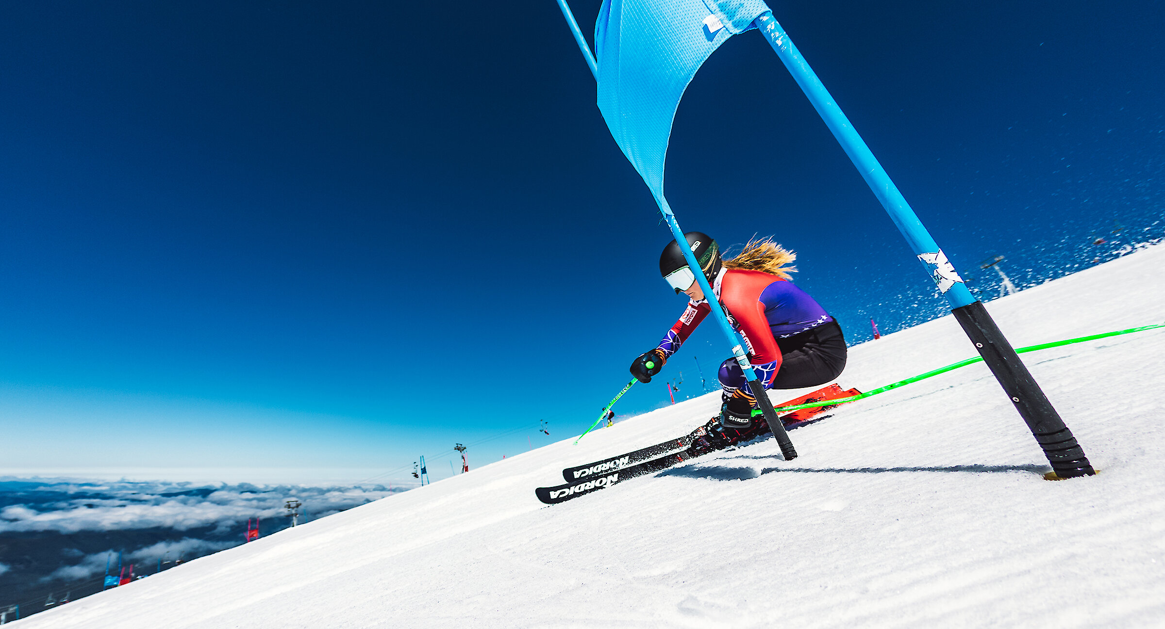Alpine ski racer carving through a slalom gate on a sunny day at Timberline Lodge, with snow spray and a deep blue sky on Mount Hood.