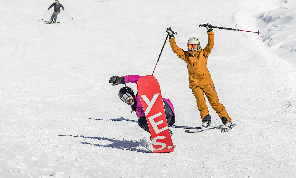 Snowboarder carving downhill while a skier celebrates behind them on a sunny slope at Timberline.