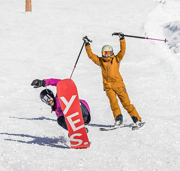 Snowboarder carving downhill while a skier celebrates behind them on a sunny slope at Timberline.