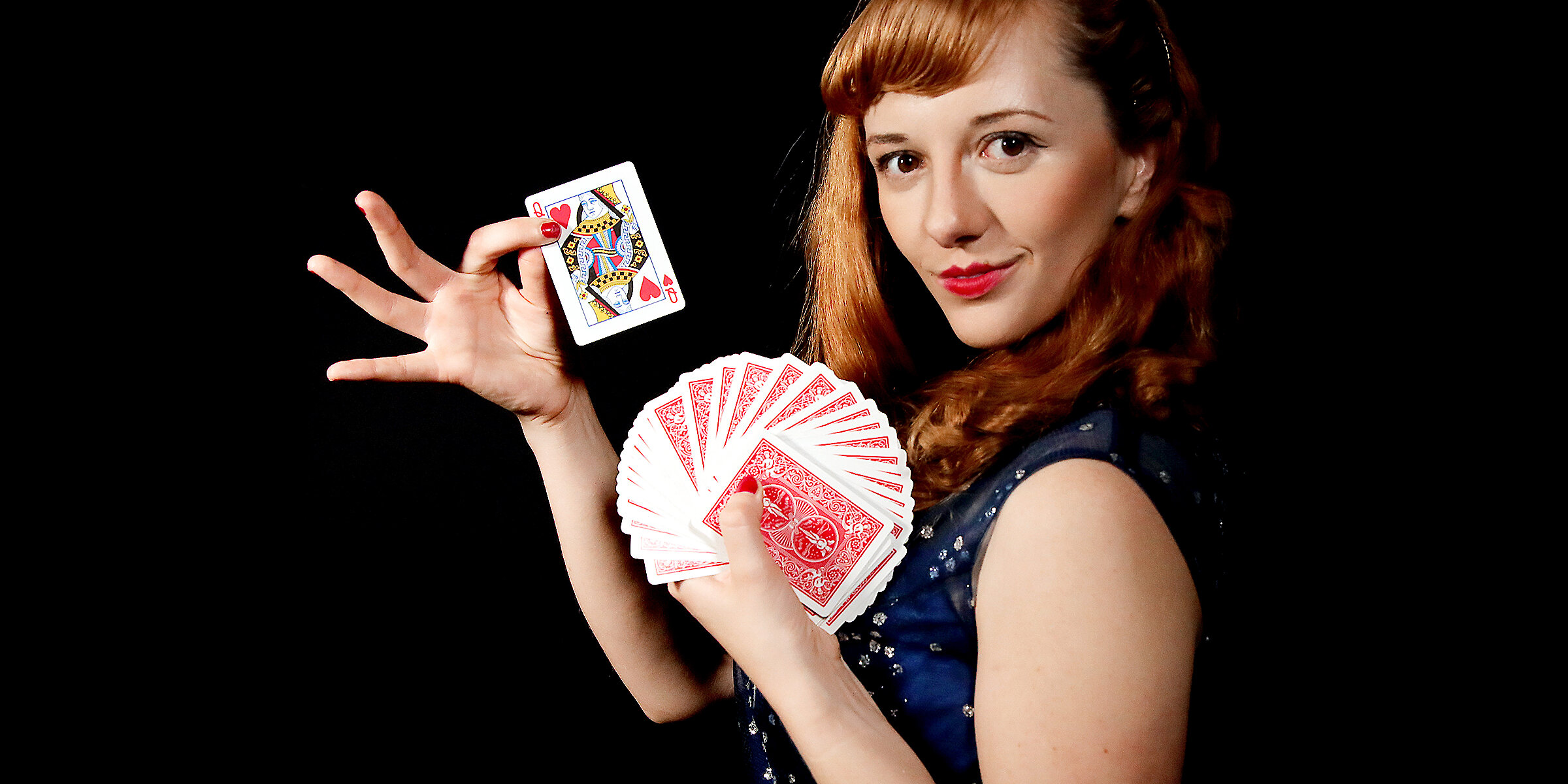 Magical Katrina, a female magician, holds a fan of playing cards and displays the queen of hearts against a black background.