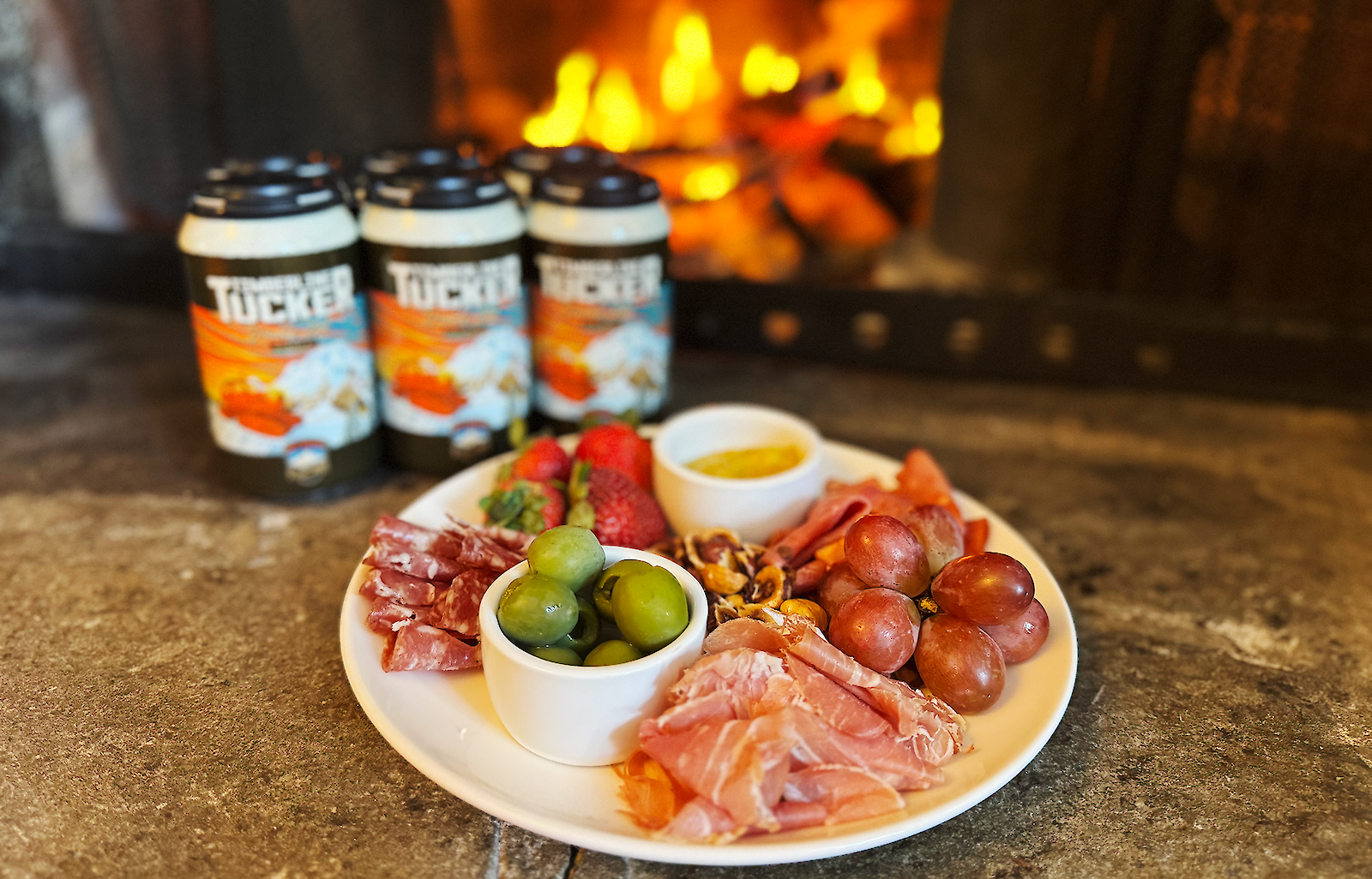 Six pack of Mt. Hood Brewing Co. Timberline Tucker Double IPA and a charcuterie plate placed in front of a roaring fire.