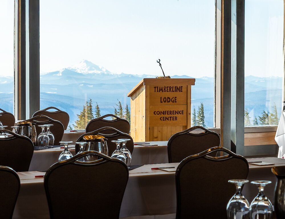 Wooden podium with "Timberline Lodge Conference Center" carved into its front pictured in front of a window featuring views of Mt. Jefferson, facing tables and chairs set up for a meeting