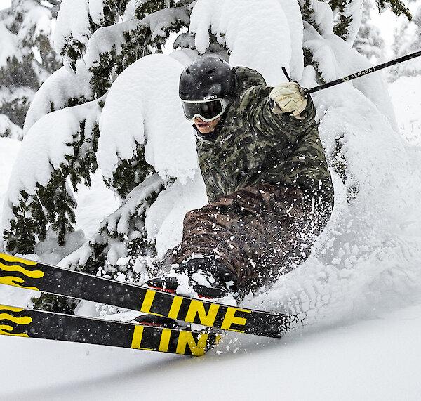 Skier in camouflage jacket carving through deep powder snow near snow-covered trees.