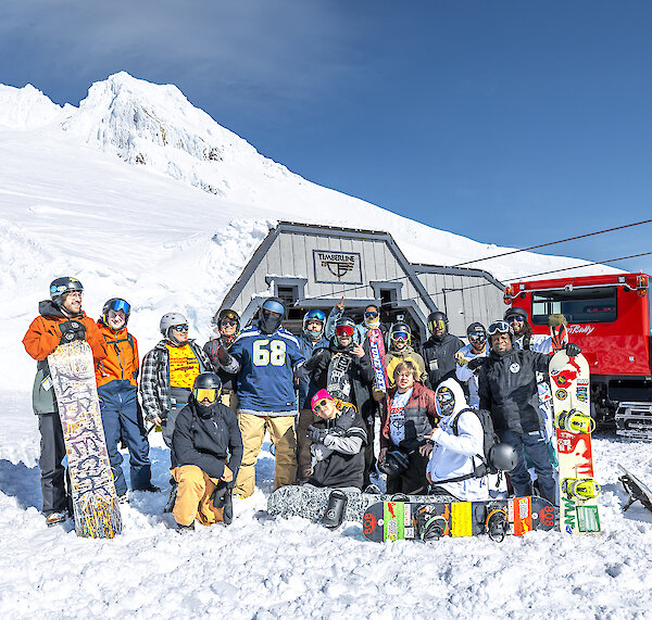 Group of skiers and snowboarders posing at Timberline on Mt. Hood in front of a snowcat and Palmer chairlift