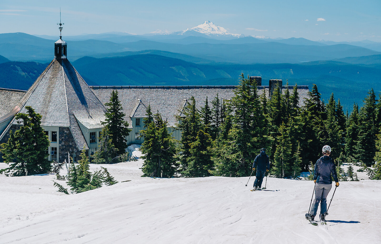 Two skiers skiing towards Timberline Lodge with Mt. Jefferson in the background
