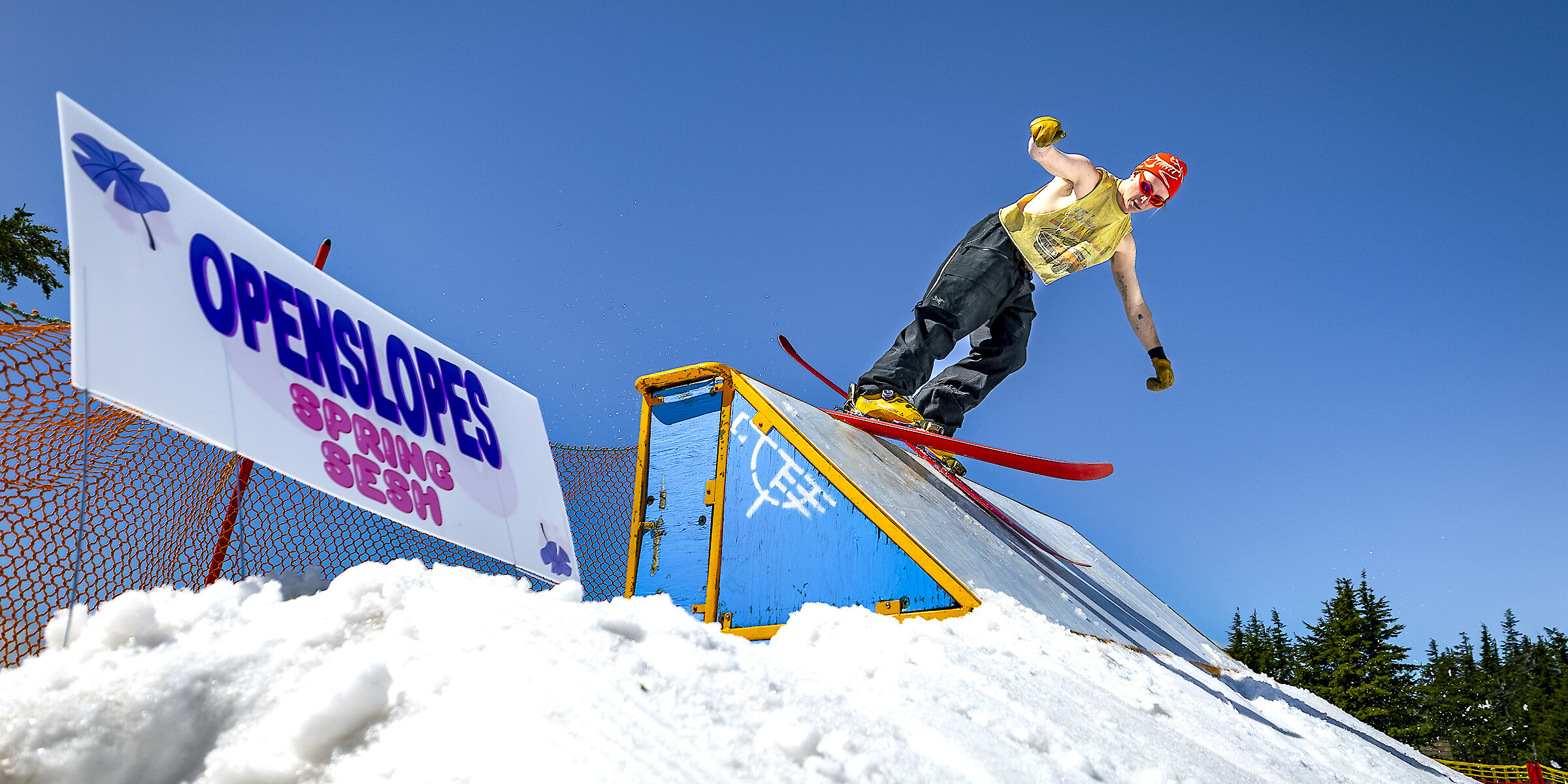 Skier performing a rail trick at OpenSlopes Spring Sesh terrain park on a sunny day with blue sky.