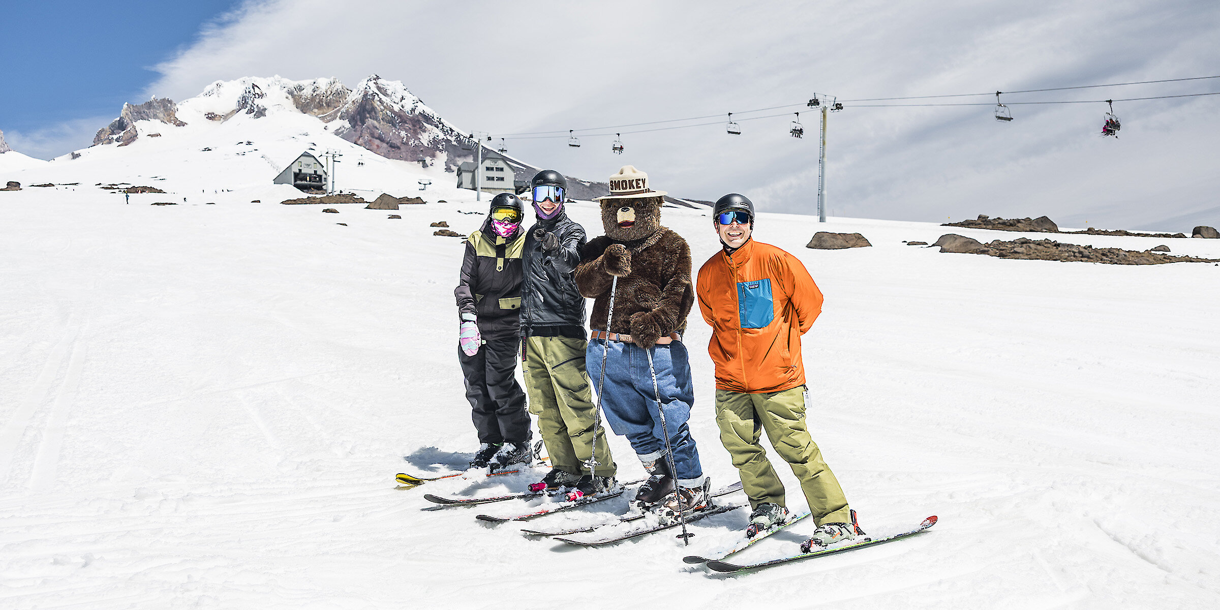 Smokey Bear skiing with three people on a snowy slope at Timberline Lodge, with Mount Hood and chairlifts in the background.