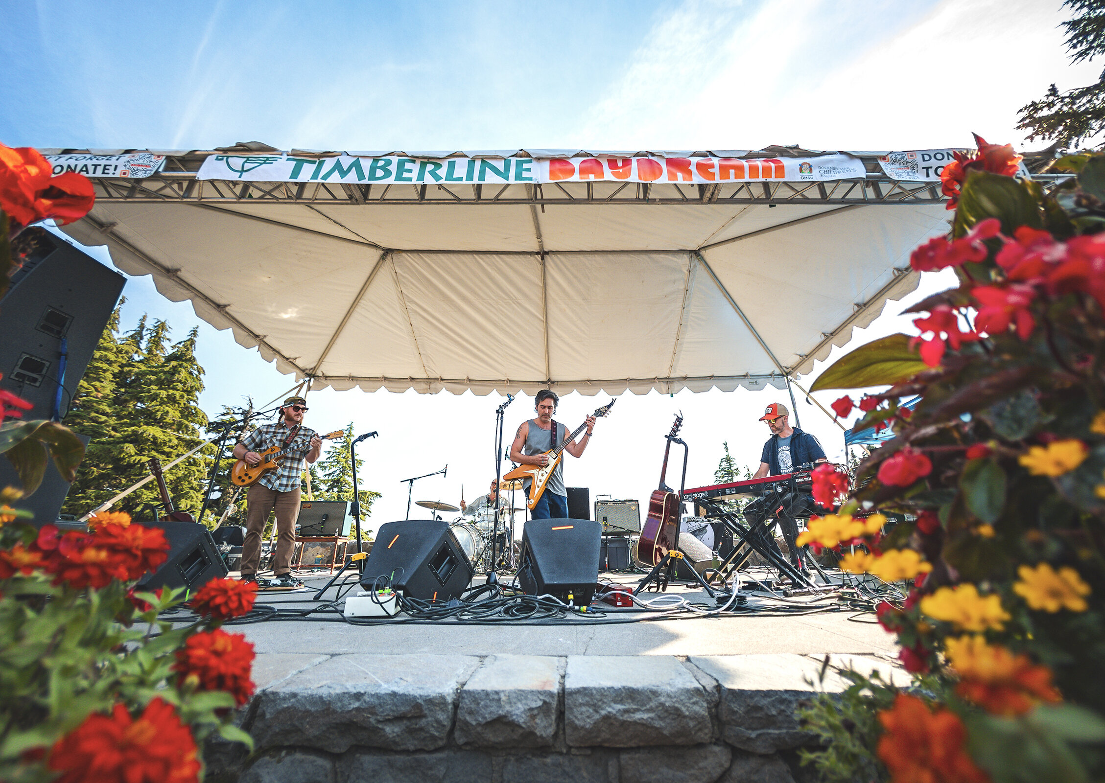 Outdoor live band performing on a stage at Timberline Lodge amphitheater, surrounded by colorful flowers under a sunny mountain sky