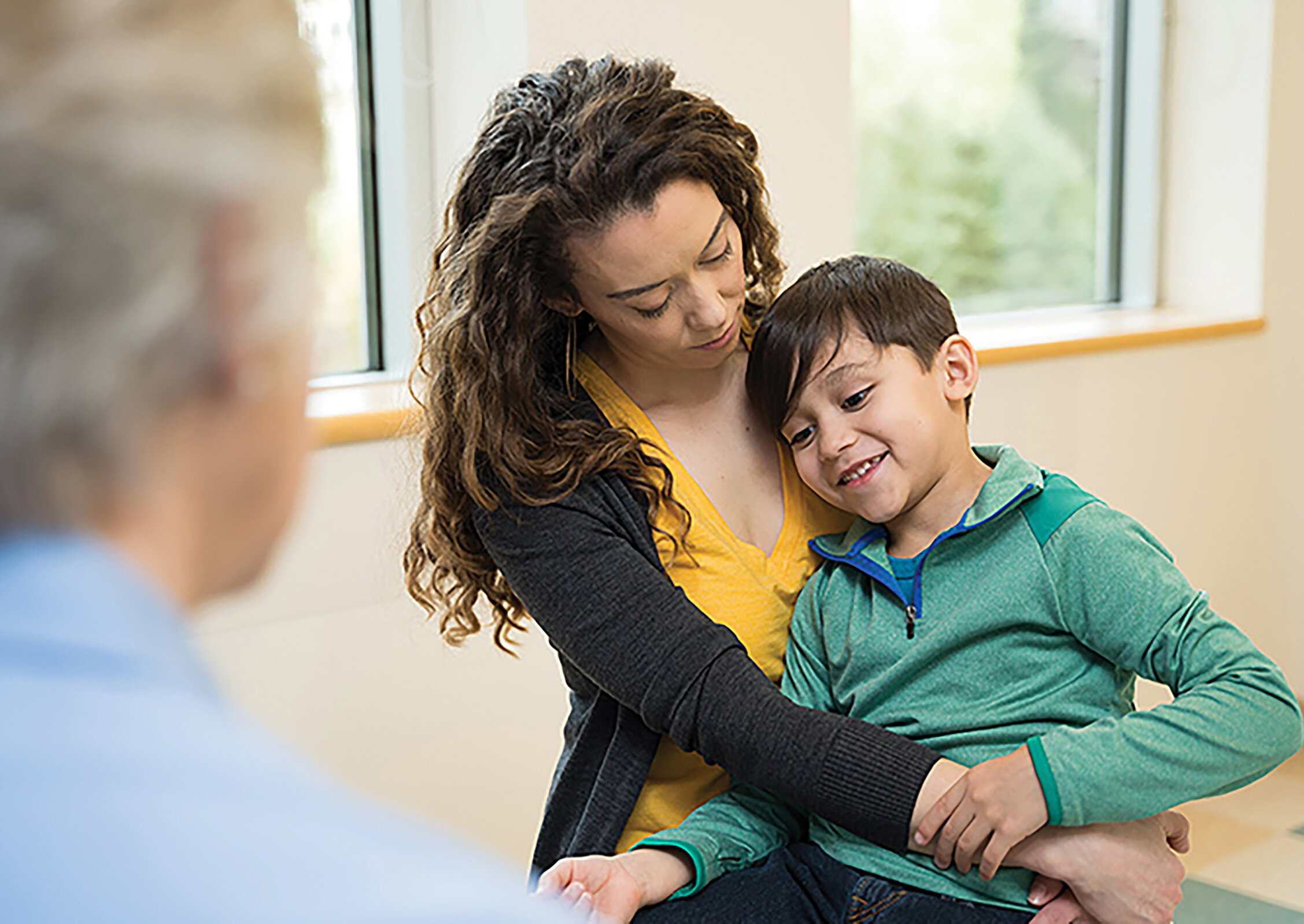 A woman gently holding and comforting a smiling young boy while seated in a medical setting, with a clinician blurred in the foreground.