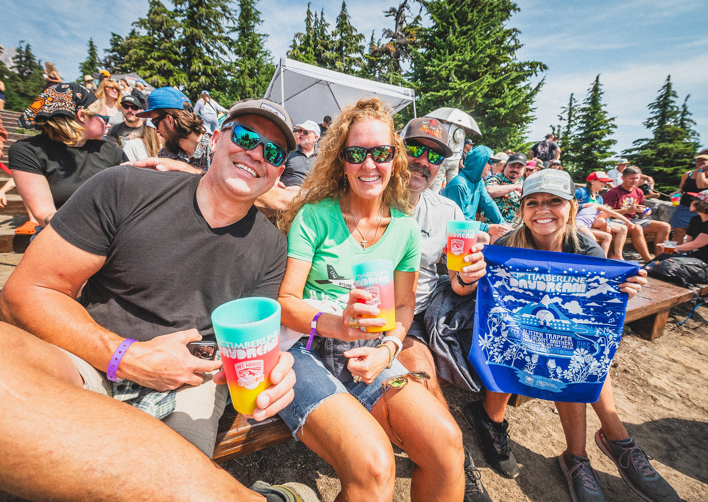 Group of friends smiling on outdoor bleachers at Timberline Daydream, holding drinks and a festival bandana, with a lively crowd and trees in the background.