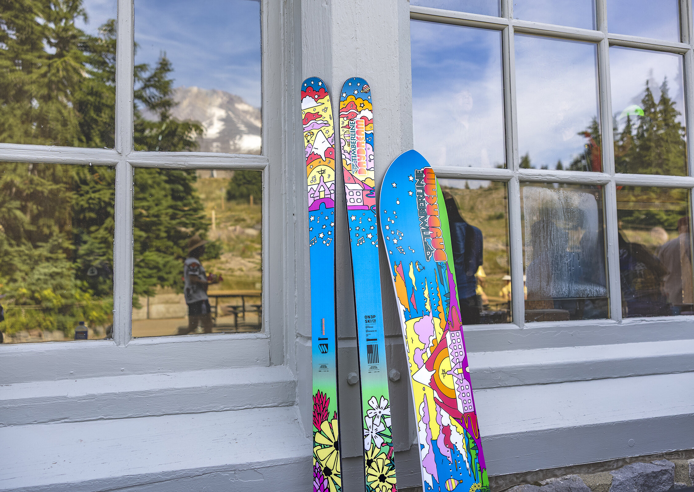Colorful Timberline Daydream skis and snowboard lean against a lodge window, reflecting trees and Mount Hood in the background.