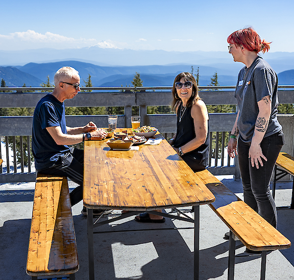 Couple and a server enjoying lunch at a picnic table on the Y'Bar patio with views of the Cascades in the background in the summer