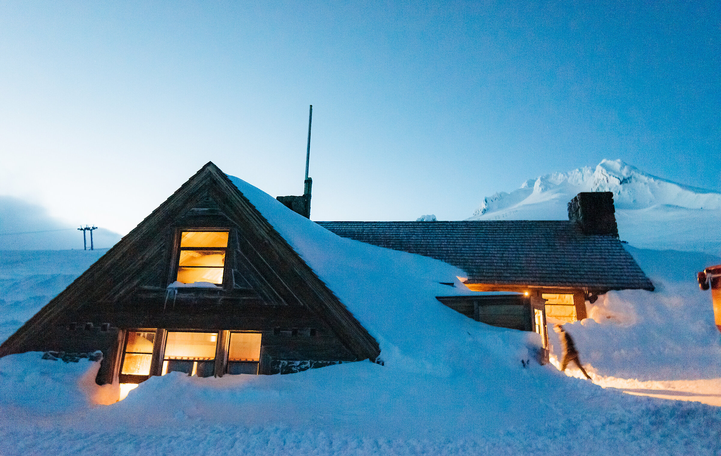 Guest entering Silcox Hut as its covered in snow at dusk, with snowy Mt. Hood pictured behind it