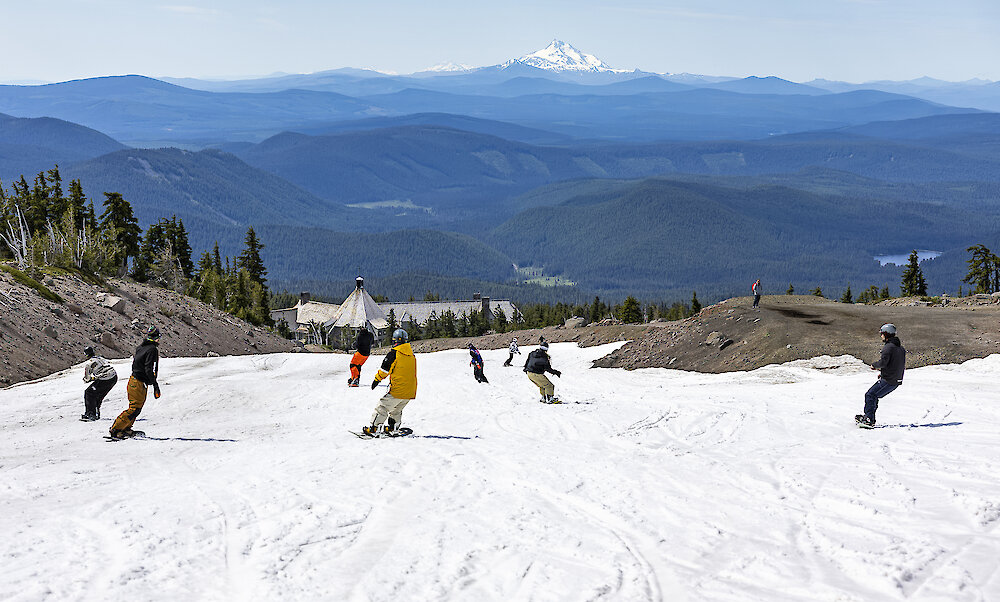 Snowboarders riding down the summer snow towards Timberline Lodge with the Cascade Range layered in the background