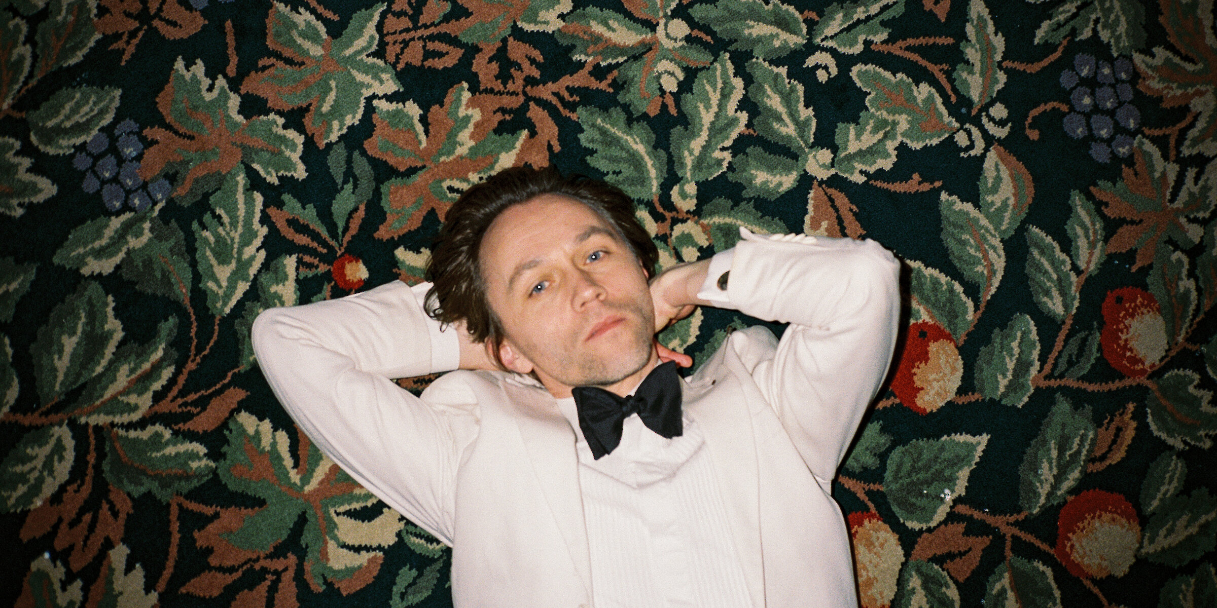 Photo of musician Sondre Lerche, pictured from above in a white tux laying on a floral carpet floor.