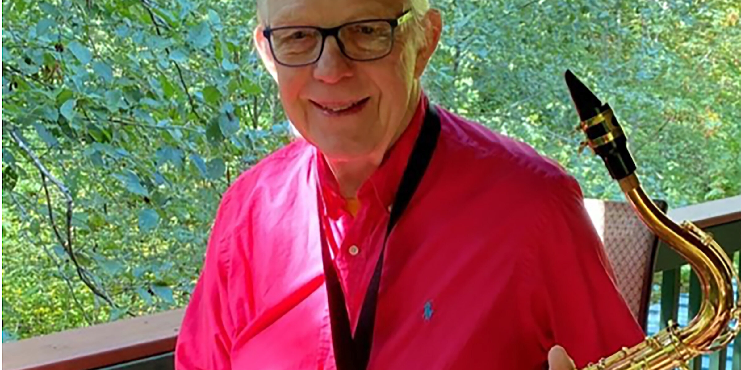 Photo of Steve Ludeman with his tenor sax, sitting on a deck with foliage in the background