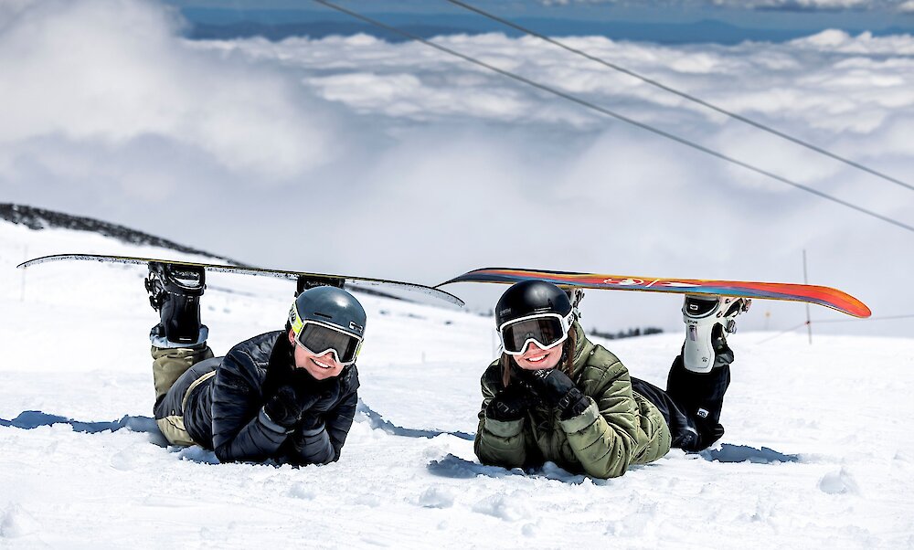 Two snowboarders lying on the snow at Timberline Lodge ski area with clouds covering the valley below.