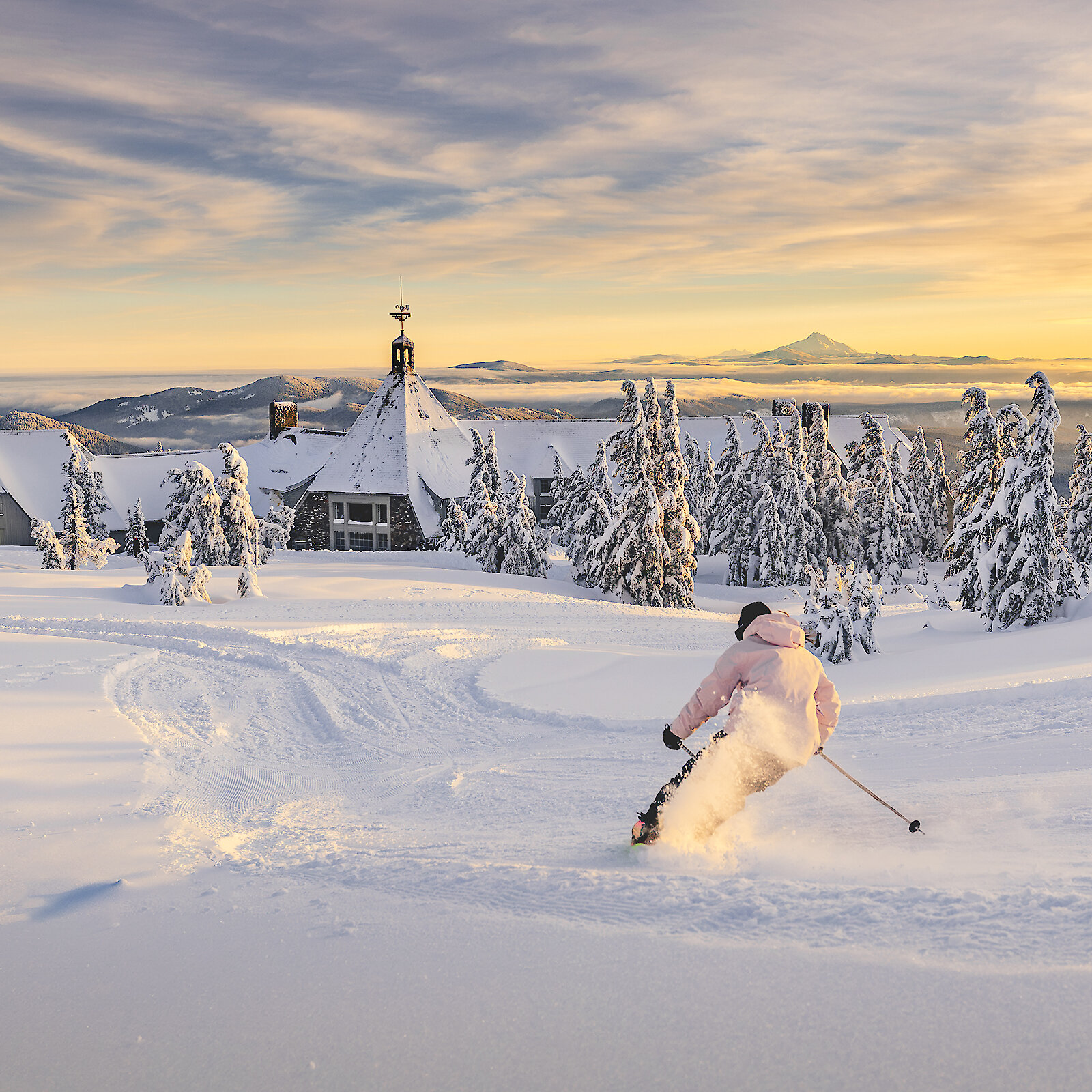 Skier skiing through the powder towards the back of Timberline Lodge during a beautiful sunset, with Mt. Jefferson in the background.