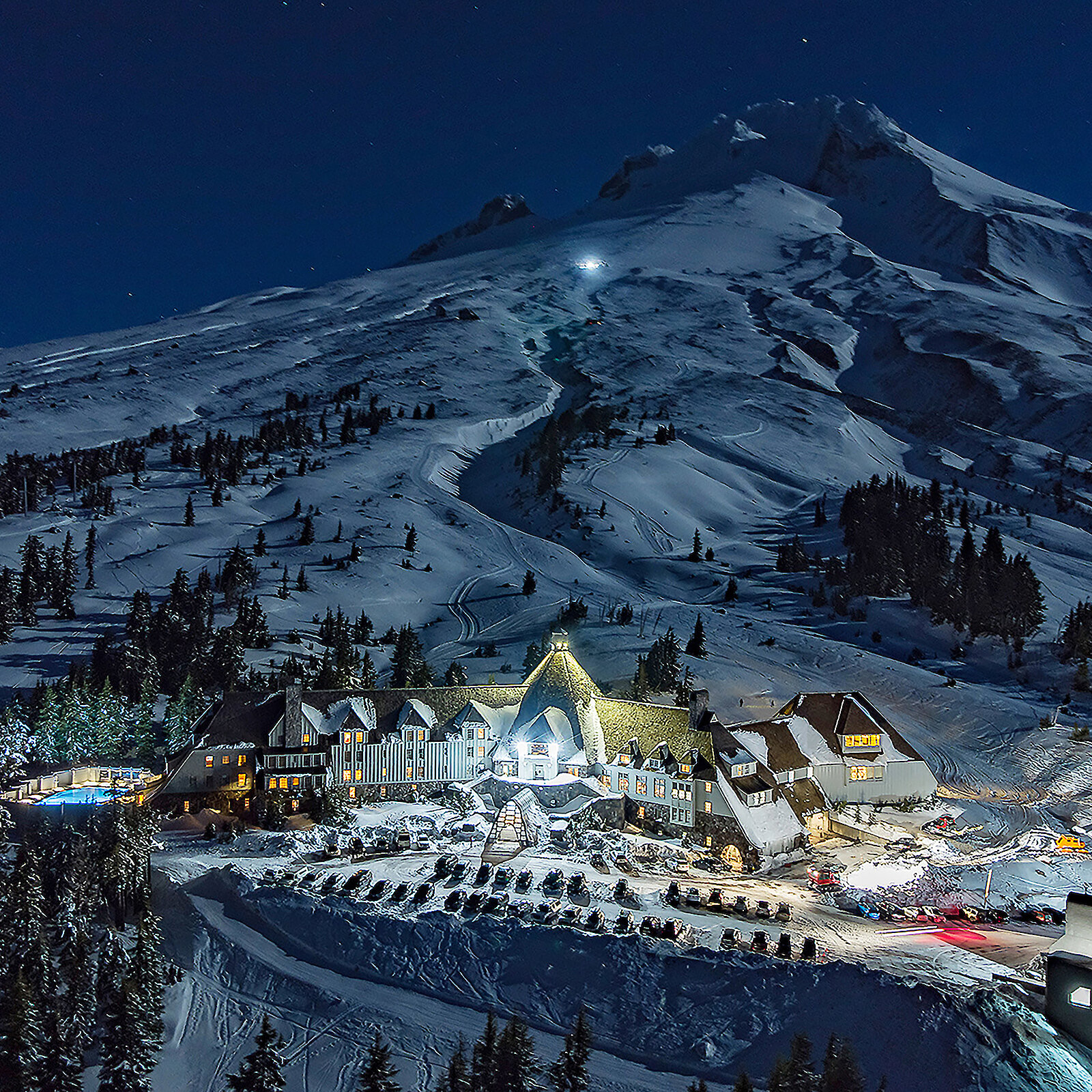 Timberline Lodge illuminated at night, surrounded by snow-covered slopes on Mount Hood under a clear, starry sky. The mountain rises steeply behind the lodge, glowing faintly in the moonlight.