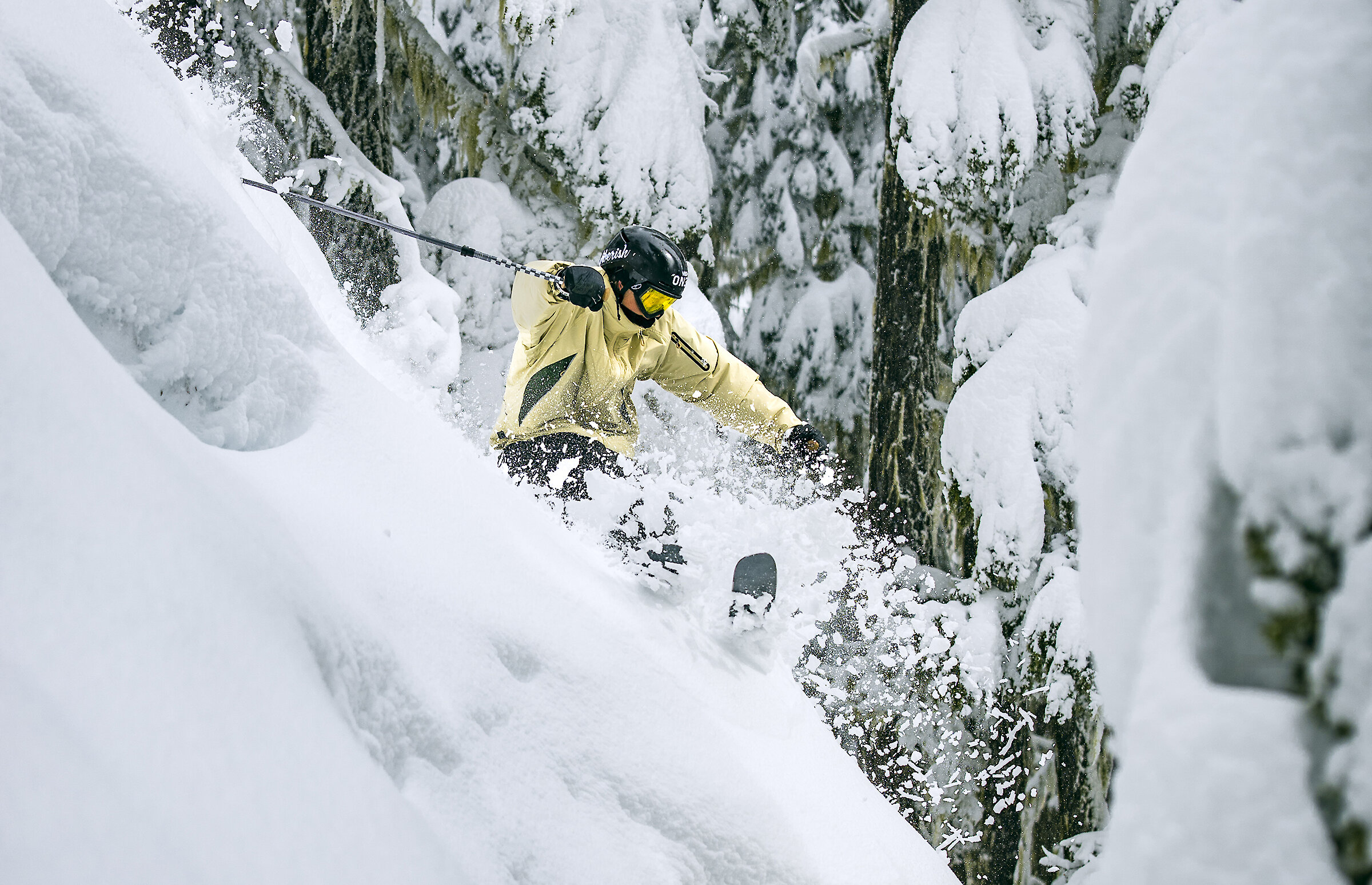 Snowboarder wearing tan gear and helmet jumps through snow-covered trees at Timberline Lodge, holding an orange snowboard midair above fresh powder.