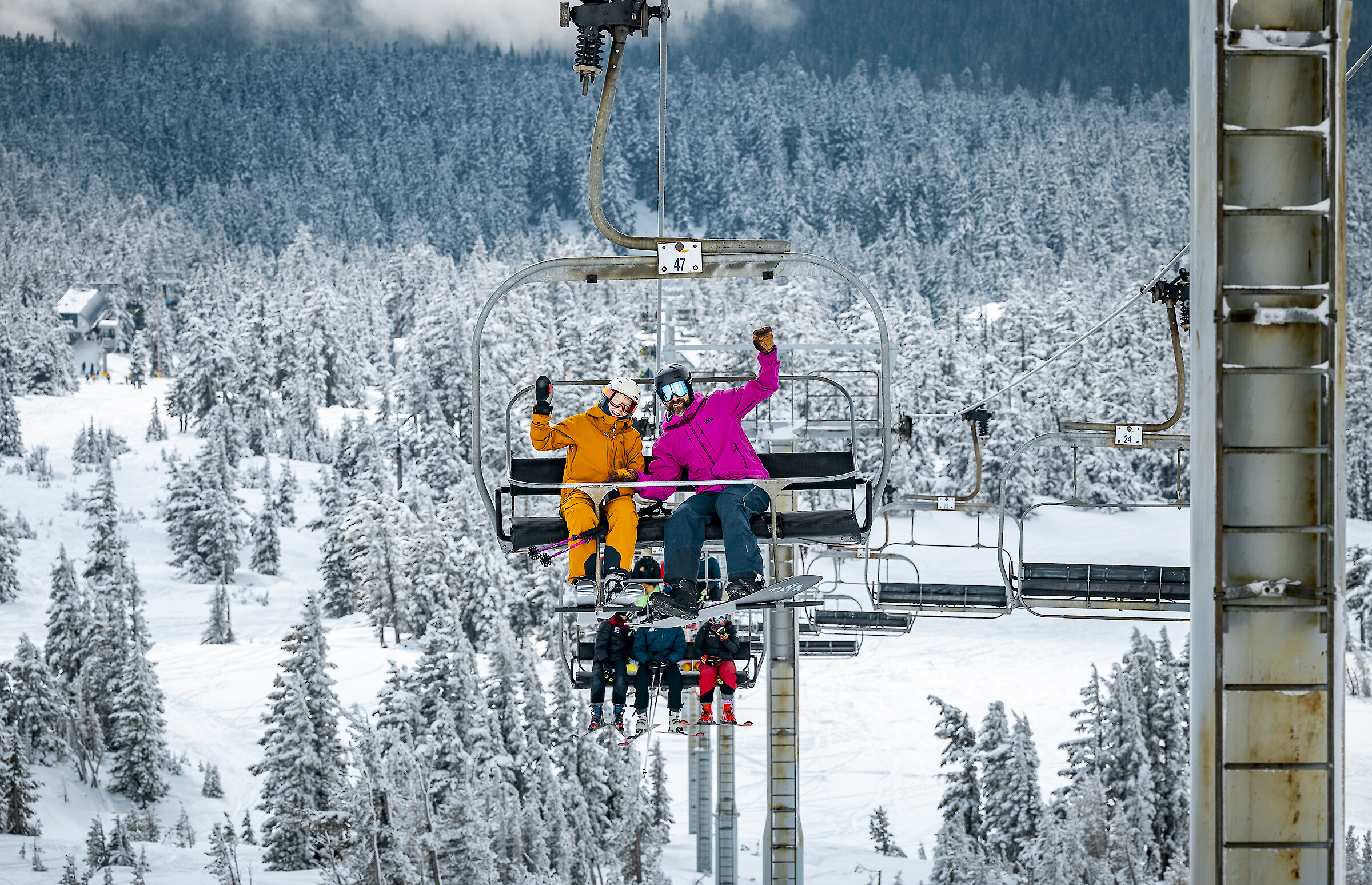 Skiers and snowboarders wave from a Timberline chairlift surrounded by snow-covered evergreen trees on Mt. Hood.