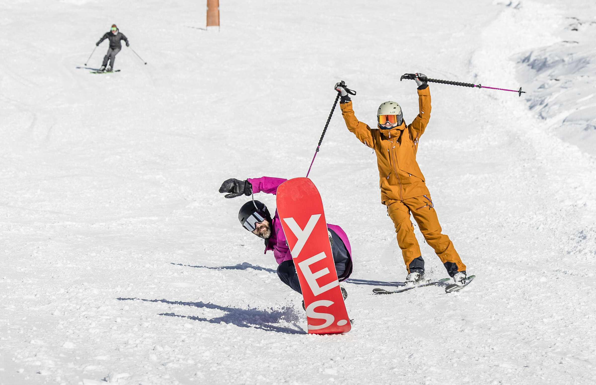 Snowboarder carving downhill while a skier celebrates behind them on a sunny slope at Timberline.