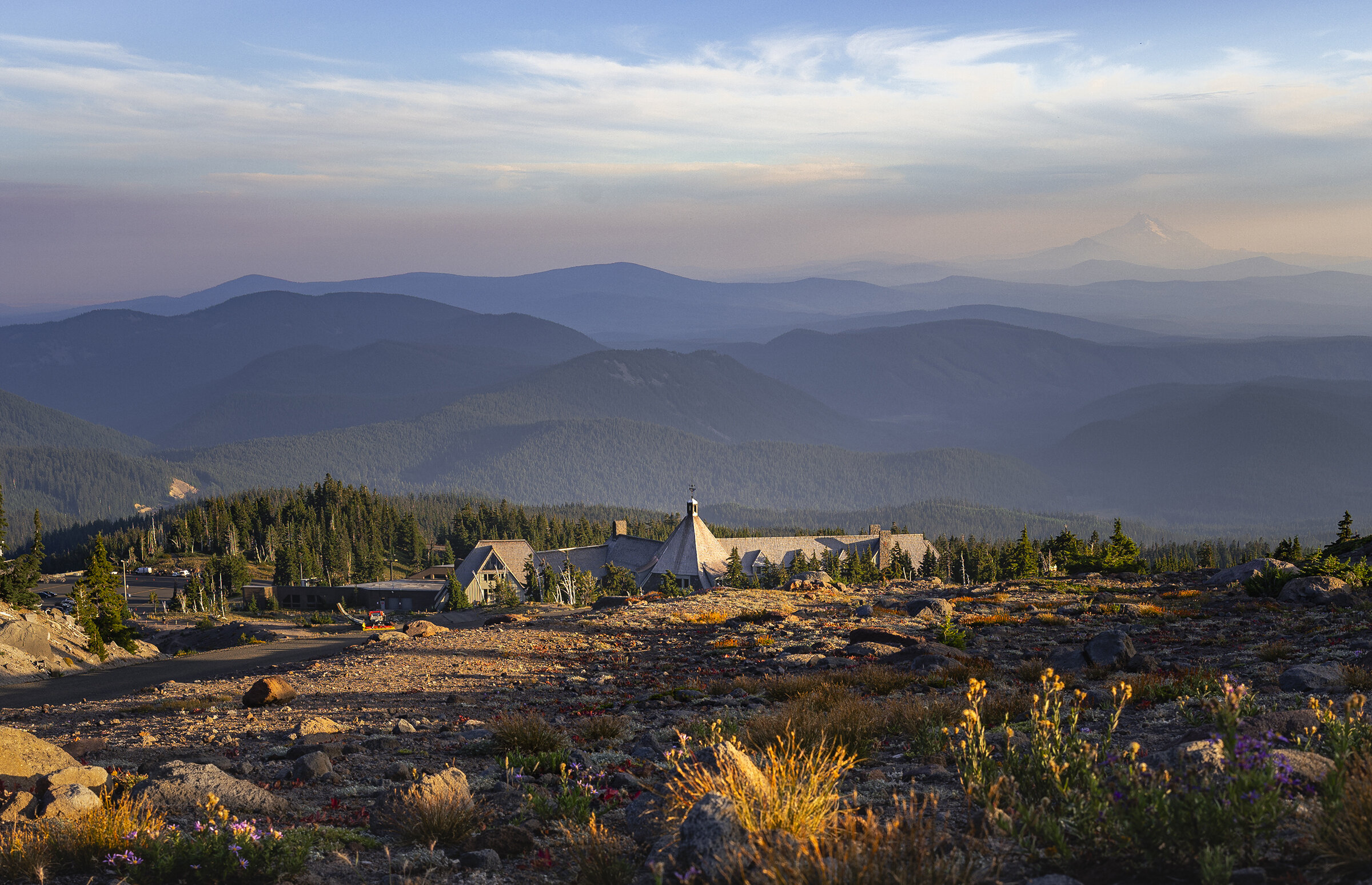 Timberline Lodge on Mount Hood surrounded by alpine wildflowers and rocky terrain, with layered Cascade mountain ridges in the distance.