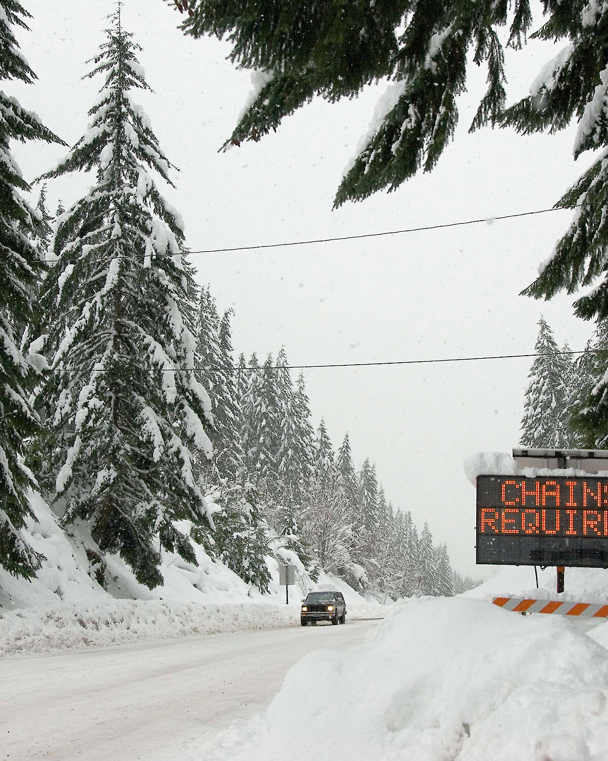 Car driving on a snow covered road surrounded by tree with a "chains required" electric sign on the right