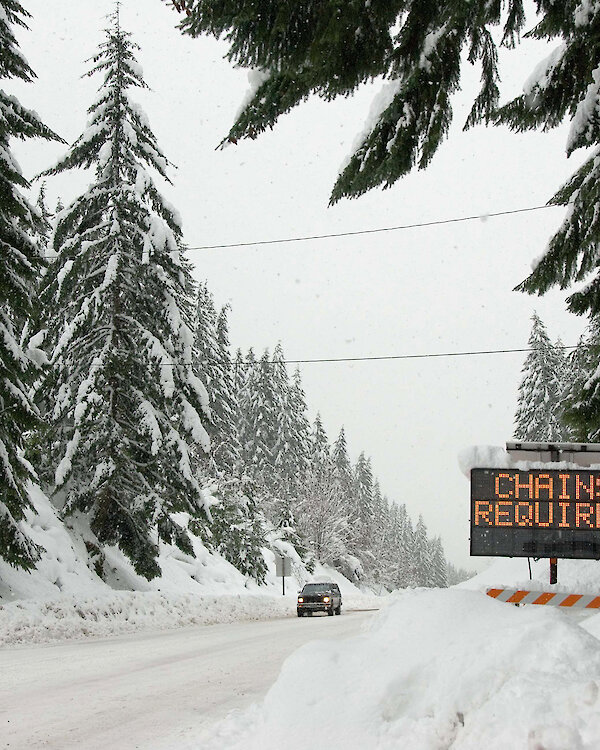 Car driving on a snow covered road surrounded by tree with a "chains required" electric sign on the right