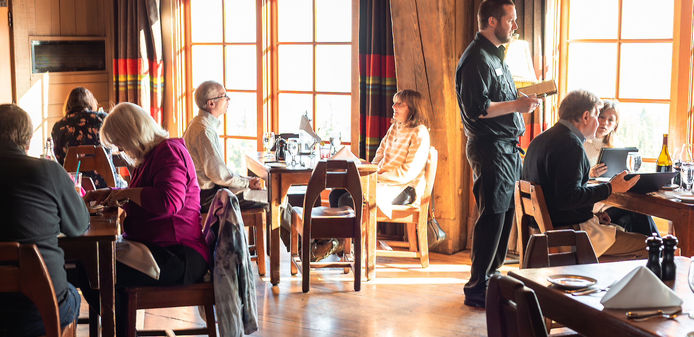 Guests enjoying lunch in the Cascade Dining Room at Timberline Lodge with a server taking orders by the sunlit windows.