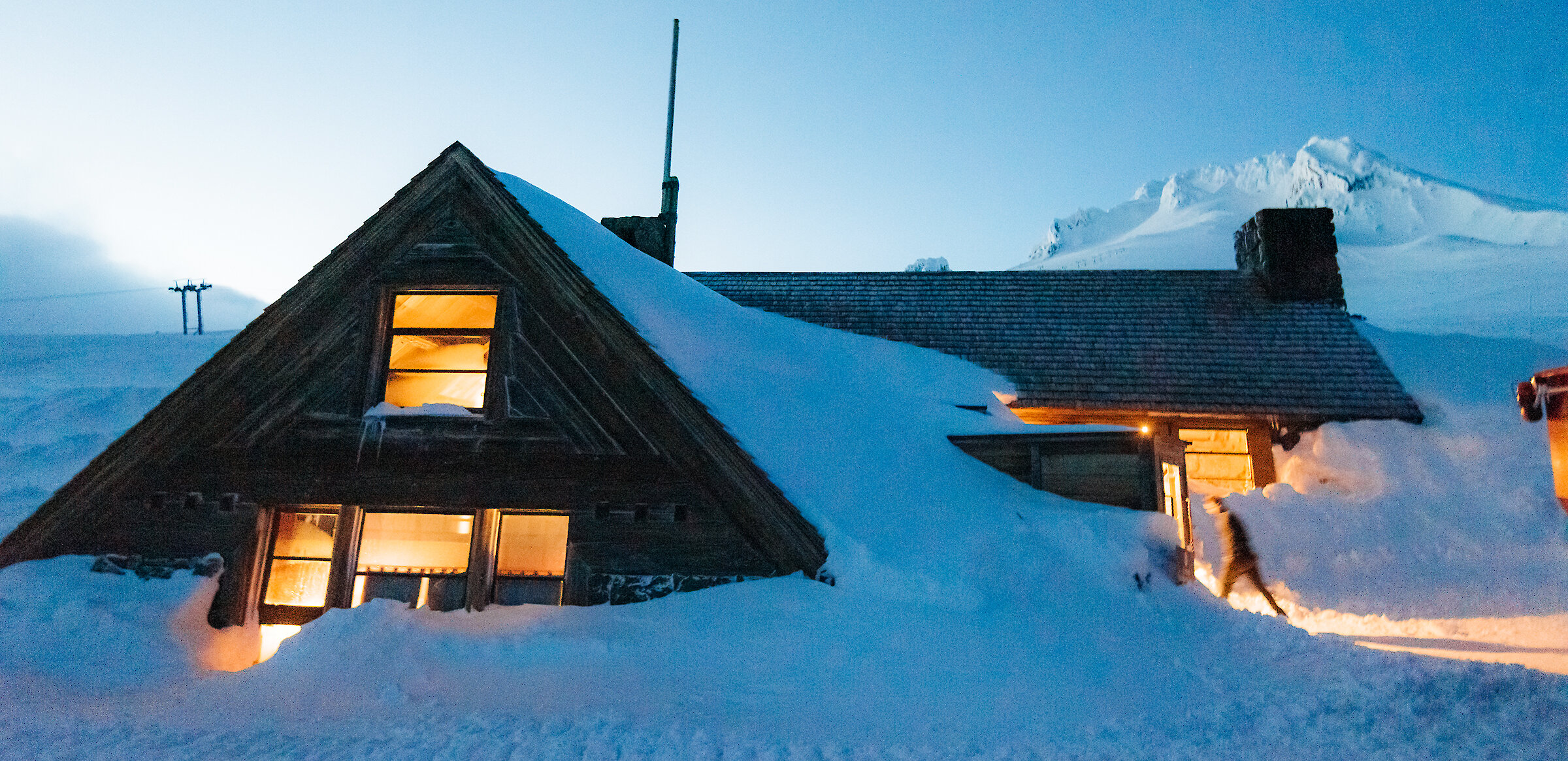 Silcox Hut pictured at dusk in the snow, with light streaming out of the windows and Mt. Hood looming in the background